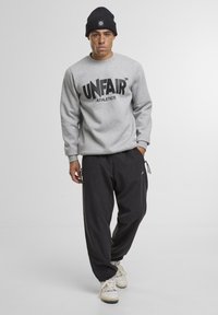 Grijze trui met "UNFAIR ATHLETICS"-opdruk, gecombineerd met zwarte joggers en witte sneakers. Model draagt een zwarte gebreide beanie. Eenvoudige, moderne stijl.