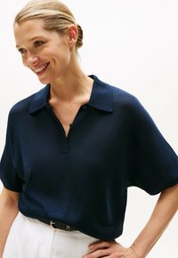 Marineblå polo t-shirt med en afslappet pasform, korte ærmer og en knappet placket. Tekstureret stof og subtil glans, parret med hvide bukser.