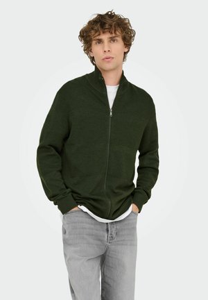 Pull à fermeture éclair vert foncé avec des motifs texturés et des poignets côtelés, porté sur une chemise blanche, associé à un jean gris clair.