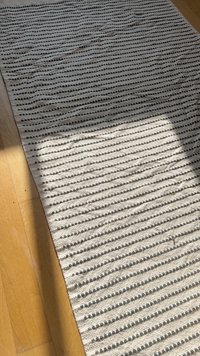 Teksturt teppe laget av vevde striper i beige og mørk grå. Materialet virker naturlig. Har et subtilt mønster og en kantet kant.