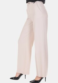 Donna che indossa pantaloni beige a vita alta e gamba larga abbinati a décolleté nere con punta affilata su sfondo bianco.