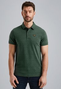 Tamnozelena polo majica s diskretnim uzorkom lišća, tri gumba uz gornji deo i malim zlatnim grbom na grudima.