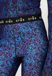 Nära bild av mönstrade leggings med ett livligt blått och lila djurtryck, med en svart midja som visar varumärket "crās."