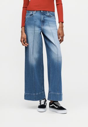 Γυναίκα που φοράει μπλε τζιν με φαρδιά μπάτζια και ραφές μπροστά, ασπρόμαυρα αθλητικά παπούτσια και μακρυμάνικο μπλουζάκι σε σκουρόχρωμη σκουριασμένη απόχρωση, στέκεται μπροστά σε απλό φόντο.