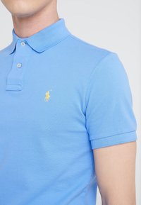 Ljusblå pikétröja i bomullsblandning, med klassisk krage, korta ärmar och en liten gul logotyp på vänster bröst.