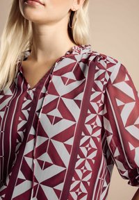 Femme blonde portant un chemisier à motifs géométriques bordeaux et blancs avec des manches bouffantes et un col à volants, accessoirisé d'une boucle d'oreille en or.