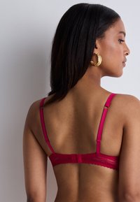 Soutien-gorge rouge avec dentelle, dotée de bretelles ajustables, d'une fermeture arrière à crochet et œillet, et d'une texture lisse, présentant un motif délicat dans le tissu.