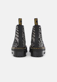 Black ankle boots with a chunky sole, featuring transparent mesh sides with a black and gray pattern, yellow accent tags, and contrasting stitching.