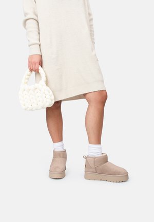 Botas de tobillo de ante beige, vestido crema acanalado, bolso trenzado blanco y calcetines blancos visibles. Enfoque en los detalles del calzado y los accesorios.