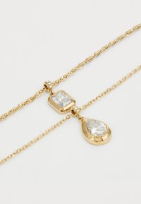 Gouden ketting met twee hangers: een vierkante en een druppelvormige, beide met heldere, gefacetteerde stenen en een gestructureerde ketting.