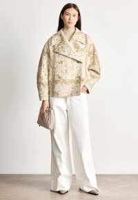 Sieviete valkā bēša krāsas teksturētu dubultpogas žaketi, baltas platajumainas bikses, bēša krāsas augstu kaklu aptverošu džemperi, rokās tur bēša krāsas maku, stāv uz bēša krāsas grīdas.