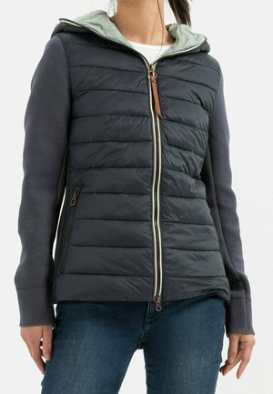 Femme portant une veste matelassée gris foncé avec capuche, fermeture éclair devant et poches latérales zippées, associée à un jean bleu.