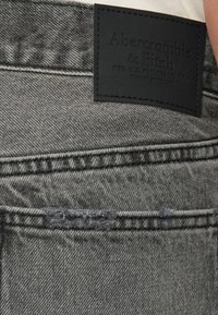 Jean en denim gris avec une surface texturée, arborant une étiquette en cuir noir cousue sur la poche arrière et des détails de couture contrastants.