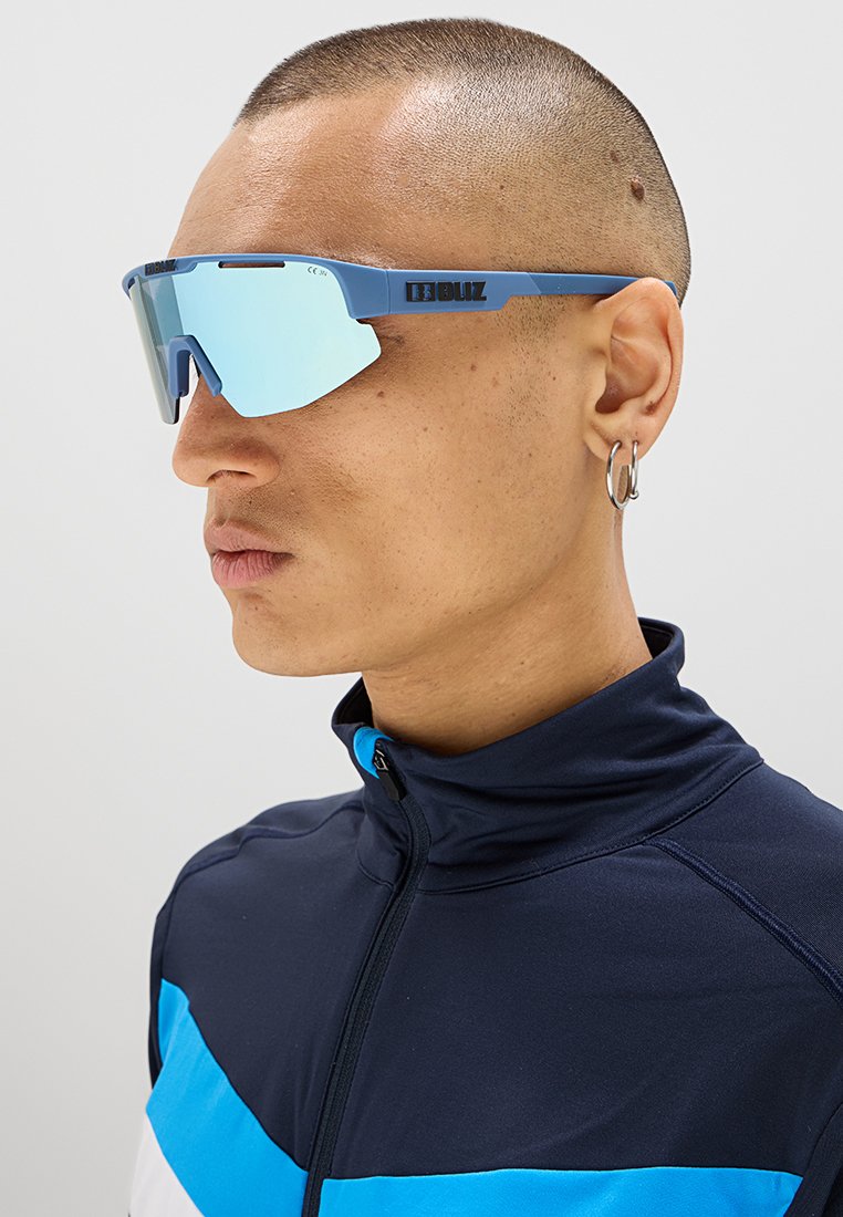 Blaue Sportsonnenbrille mit verspiegelt Glas, kantigem Rahmen und dezentem Branding. Getragen von einer Person in einer dunklen Jacke mit blauen Akzenten.