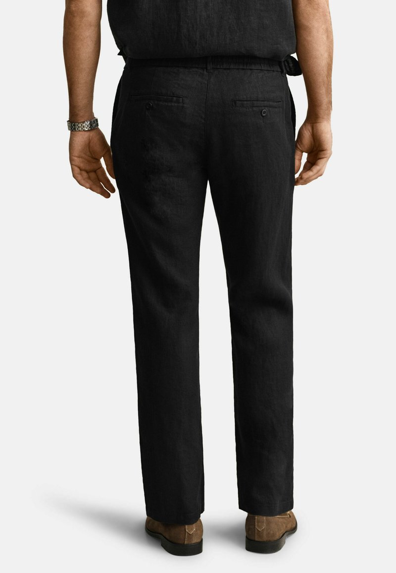 Mann trägt schwarze gerade geschnittene Hose, braune Schuhe und eine silberne Uhr, von hinten vor hellem, einfarbigem Hintergrund gezeigt.