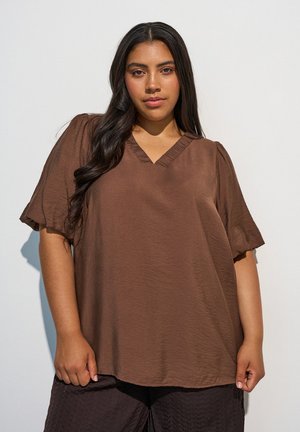 Femme aux longs cheveux foncés portant un chemisier marron ample à col en V et un pantalon texturé foncé, debout devant un fond clair.