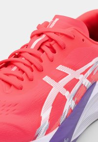 Roter Sportschuh mit strukturiertem Obermaterial, weißem gestreiftem Logo und lila Sohle. Verfügt über kontrastierende Schnürsenkel und einen gepolsterten Schaft.