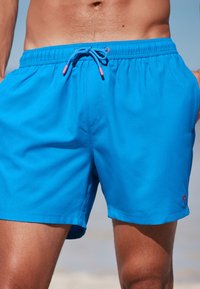 Kék úszóshort, rugalmas derékkal és zsinórral, rózsaszín kiegészítőkkel. A short sima tapintású, kis piros logóval rendelkezik.