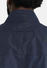 Navyblauwe jas met een hoge kraag, voorzien van een gestikt logo-label op de achterkant. De stof lijkt glad met een zachte textuur.