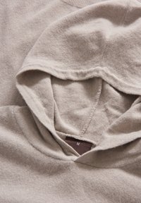 Beige Kapuzenpullover mit weicher Textur, ausgestattet mit einer Fronttasche und einer Kapuze mit Kordelzug. Etikett zeigt Größe M an.