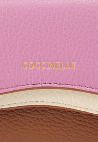 Ροζ δερμάτινο πορτοφόλι με χρυσό λογότυπο "Coccinelle". Διαθέτει υφή επιφάνειας, λευκές και καφέ λεπτομέρειες, καθώς και ραφές με αντιθετικό χρώμα στα άκρα.