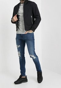 Cazadora negra con cuello alzado, camiseta gris debajo, jeans azules desgastados y zapatillas negras. Tatuajes visibles en los antebrazos.