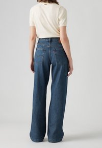 Mörkblå denim byxor med vida ben och hög midja, med bakfickor och en brun läderlogga på midjebandet.