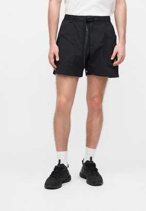 Mees kannab musti elastse vöökohaga lühikesi pükse esitaskutega, valgeid sokke, musti spordijalatsid ja valget särki, seisab taustal ilma mustrita.