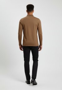 Mann, der mit dem Rücken zur Kamera steht, trägt ein braunes Langarmshirt, schwarze Hose und schwarze Schuhe vor einem schlichten weißen Hintergrund.
