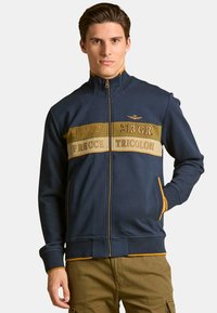 Chaqueta de punto azul marina con bandas contrastantes en beige y dorado, que presenta texto, un pequeño emblema y bolsillos laterales. Tejido suave y puños acanalados.