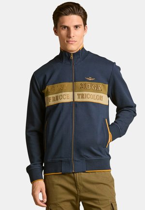 Giacca zip-up blu navy con bande a contrasto beige e dorate, dotata di testo, un piccolo emblema e tasche laterali. Tessuto morbido e polsini a coste.