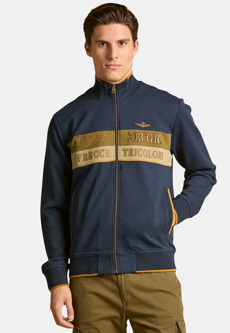 Chaqueta de punto azul marina con bandas contrastantes en beige y dorado, que presenta texto, un pequeño emblema y bolsillos laterales. Tejido suave y puños acanalados.