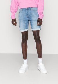 Shorts in denim azzurro con orli risvoltati, abbinati a una felpa rosa e sneakers bianche, che presentano un look casual e rilassato.
