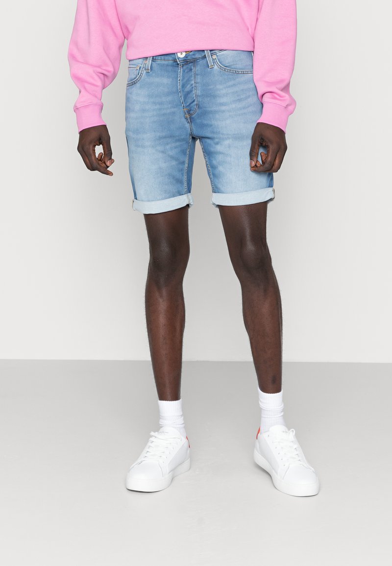 Shorts in denim azzurro con orli risvoltati, abbinati a una felpa rosa e sneakers bianche, che presentano un look casual e rilassato.