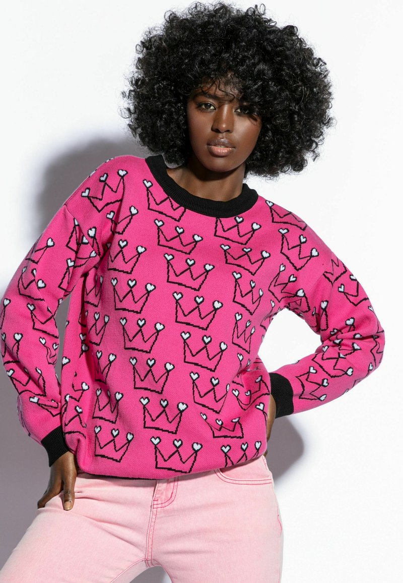 Różowy sweter z czarnymi ściągaczami przy mankietach i dekolcie, ozdobiony powtarzającym się wzorem koron w czarnym obrysie oraz białymi akcentami w kształcie serc.