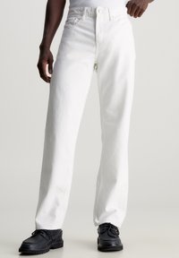 White denim jeans with a straight-leg cut, featuring a button and zipper closure, front pockets, and a textured fabric finish. Black shoes worn.