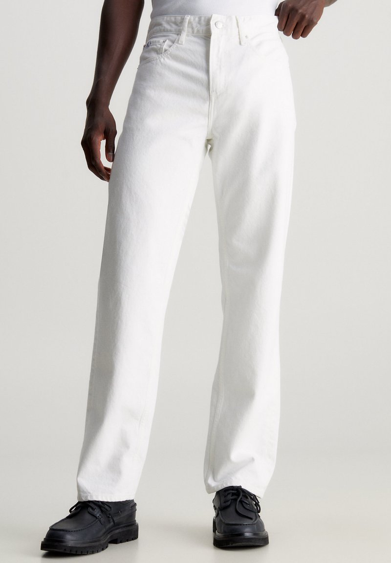 White denim jeans with a straight-leg cut, featuring a button and zipper closure, front pockets, and a textured fabric finish. Black shoes worn.