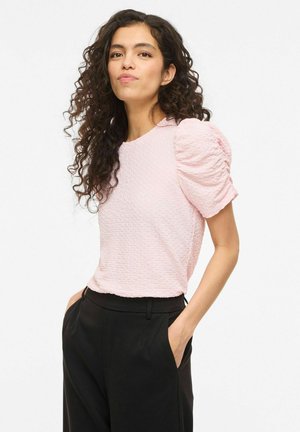 Kobieta o długich kręconych włosach, ubrana w jasnoróżowy, fakturowany top z bufkami i czarne spodnie, stoi z rękami w kieszeniach na białym tle.