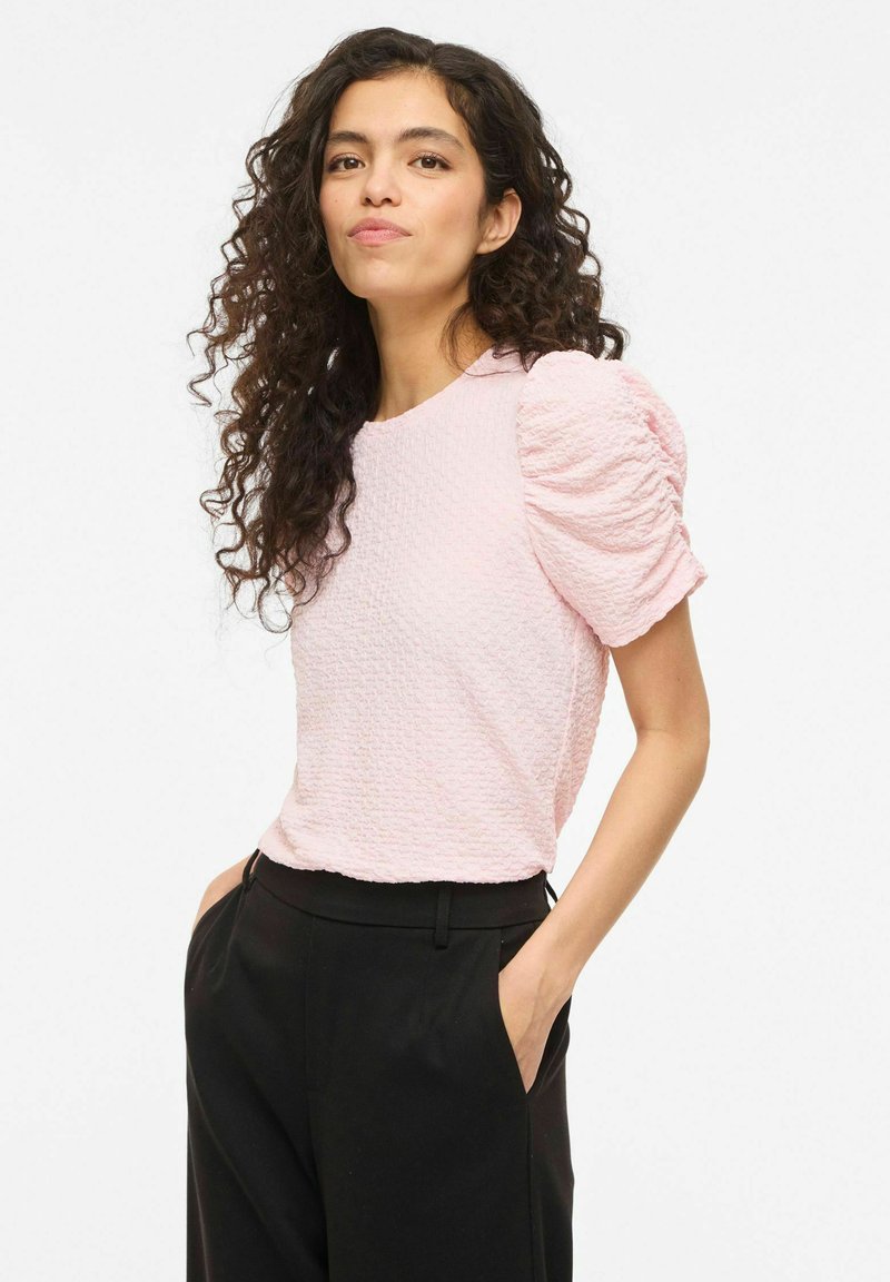 Žena s dlhými kučeravými vlasmi má na sebe textúrovaný svetloružový top s naberanými rukávmi a čierne nohavice, stojí so založenými rukami v kapse na bielom pozadí.