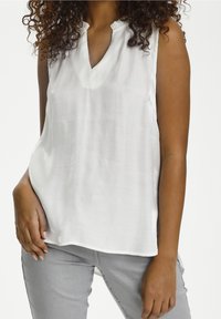 Biała bezrękawnikowa bluzka z dekoltem w serek, o miękkiej fakturze i marszczonym wykończeniu dekoltu. W połączeniu z jasnoszarymi spodniami. Prosty, minimalistyczny design.