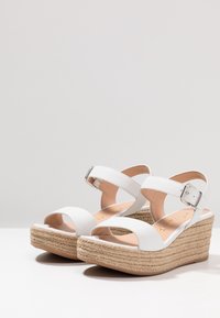 Valkoiset viistokorkoiset sandaalit, joissa on tekstuuripinta, kaksi leveää hihnaa ja solki. Pohja on tehty kudotusta juutista, mikä tarjoaa korkeutta ja vakautta.