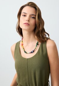 Collier de perles multicolores avec des tailles et des formes variées, y compris des perles rondes et cylindriques en vert, orange, violet et jaune.