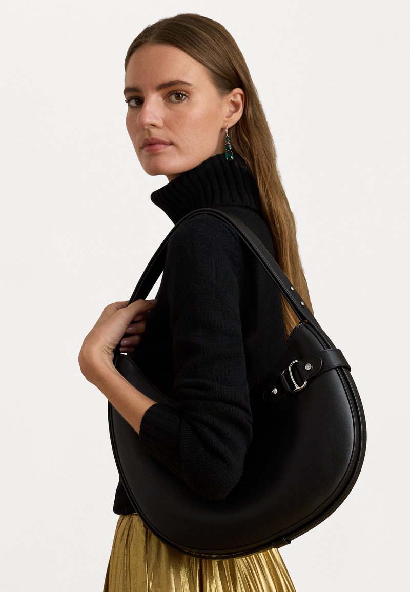 Bolsa de ombro em couro preto com formato de meia-lua, detalhes em fivela de metal prateado e uma textura suave, acompanhada de um suéter preto.