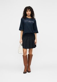 Μπλε κοντομάνικο πουκάμισο με λευκό λογότυπο, μαύρη πλισέ φούστα, καφέ δερμάτινες μπότες μέχρι το γόνατο και μικρή λευκή τσάντα χειρός. Απλός σχεδιασμός.