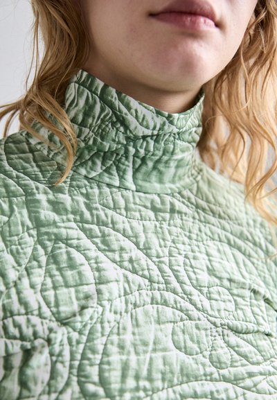 Quilted green high-neck top featuring a textured pattern and a subtle sheen, with a close-fitting collar and soft fabric finish.