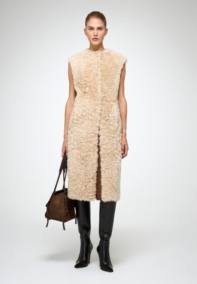 Cappotto senza maniche in shearling beige con apertura frontale, abbinato a stivali in pelle neri a gamba stretta fino al ginocchio e una borsa marrone strutturata.