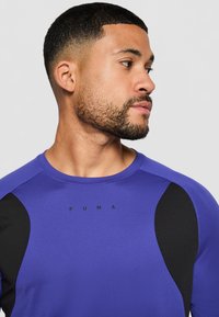 Lilla treningsskjorte med svarte detaljer, rund hals og kort erm. Har "PUMA"-logoen på forsiden. Glatt, tettsittende tekstur.