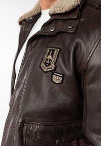 Bruine leren jas met een schapenvachtkraag, decoratieve patches en drukknoopsluiting. Geweven afwerking en twee voorzakken.