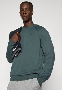 Sudadera verde de Nike con cuello redondo y mangas raglán. Toalla oscura caída sobre el hombro. Pantalones grises visibles. Diseño simple, sin patrones.