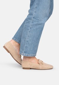 Mocasines de ante beige con tacón plano, que cuentan con un detalle decorativo de hebilla dorada y puntera redonda, combinados con unos jeans de longitud hasta el tobillo de color azul claro.